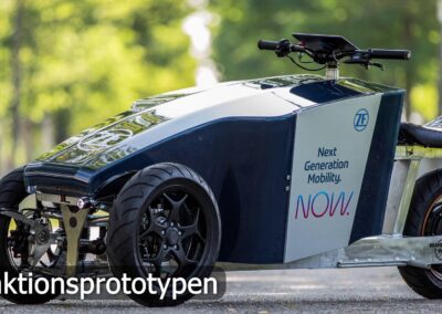 Das Bild zeigt einen innovativen Funktionsprototypen eines dreirädrigen Elektromotorrads, das im Freien auf einer asphaltierten Fläche abgestellt ist. Die Fahrzeugfront ist futuristisch und kantig gestaltet, mit zwei Vorderrädern, die über eine ausgeklügelte Aufhängung miteinander verbunden sind. Auf der Seitenverkleidung ist das Logo von ZF Friedrichshafen zu sehen, daneben der Slogan: „Next Generation Mobility. NOW.“ Das Fahrzeug verfügt über einen einzelnen Sitz mit einem gepolsterten Bezug und einen Lenker mit Displayeinheit. Der Prototyp dient wahrscheinlich der Erprobung neuer Mobilitätskonzepte und -technologien. Unten links im Bild ist der Text „Funktionsprototypen“ eingeblendet. Im Hintergrund sind unscharfe grüne Bäume zu sehen, was dem Bild eine natürliche, moderne Atmosphäre verleiht.