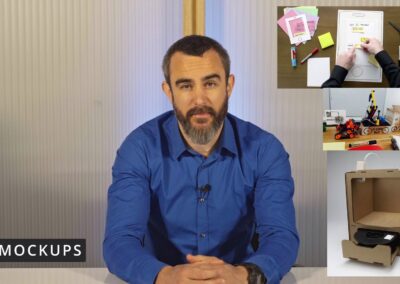 **Bildbeschreibung (barrierefrei und beschreibend):** Das Bild zeigt **Lars Laumeyer** in einem Video-Setting. Er sitzt an einem Tisch, trägt ein blaues Hemd und blickt ruhig in die Kamera. Auf seiner linken Seite (rechts im Bild) sind drei kleinere Bilder montiert, die verschiedene Arten von **Mockups** zeigen: 1. Oben: Ein **Papier-Mockup** einer Smartphone-App mit beschrifteten Zetteln, Stiften und Schere – eine klassische Methode aus dem UX-/UI-Design. 2. Mitte: Ein **funktionales Modell** aus Alltagsmaterialien mit dem Schriftzug „Klean Bot“, das ein kindlich gestaltetes Roboterfahrzeug zeigt – vermutlich ein früher Prototyp für ein Technikprodukt. 3. Unten: Ein **Mockup aus Pappe**, das einen 3D-Drucker oder ein technisches Gerät darstellt, mit aufgeklebten Bedienelementen und Einschüben – eine visuelle und räumliche Darstellung einer Produktidee. Unten links im Bild ist ein schwarzer Kasten mit weißer Schrift eingeblendet: **„MOCKUPS“** Das Bild ist Teil eines E-Learning-Videos oder Vortrags über frühe Prototyping-Methoden und Visualisierung von Ideen.