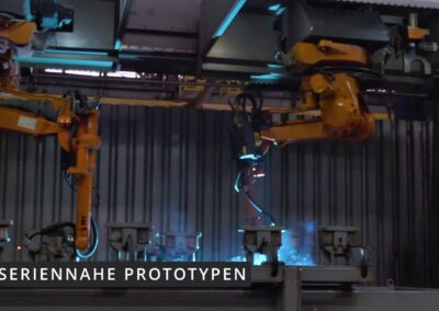 Das Bild zeigt eine industrielle Fertigungsumgebung mit zwei orangefarbenen Roboterarmen, die an einem metallischen Gestell montiert sind. Die Roboter führen automatisierte Schweißarbeiten an Werkstücken durch, die in Vorrichtungen eingespannt sind. Blaues Licht und Funkenflug deuten auf laufende Schweißprozesse hin. Im Hintergrund ist eine dunkle Metallwand zu sehen, die die Werkhalle begrenzt. Die Szene ist in ein kühles, technisches Licht getaucht. Unten links im Bild steht in weißer Schrift auf schwarzem Hintergrund der Text: „Seriennahe Prototypen“ – ein Hinweis darauf, dass hier Prototypen getestet oder gefertigt werden, die bereits stark an das spätere Serienprodukt angelehnt sind.